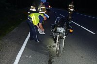 Na fotografii widać drogę, na której znajduje się rozbity motocykl. Z boku motocykla widać pochylonego policjanta. Obok policjanta widać trzech  strażaków, a w tle fotografii widać wóz strażacki.