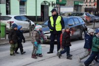 Na fotografii widać przejście dla pieszych (zebra), po którym przechodzą dzieci. Pośród nich znajduje się policjant, który wstrzymał ruch pojazdów.