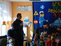 Na fotografii widać policjanta, który w dłoni trzyma mikrofon. Z prawej strony zdjęcia widać grupę dzieci.