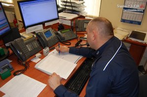 Na zdjęciu widać siedzącego przy biurku policjanta, który rozmawia przez telefon stacjonarny. Na biurku widać monitory komputerowe oraz telefony i radiostacje.