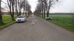 na zdjęciu fragment drogi, po lewej srebrny samochód, po prawej przy drzewie inny rozbity pojazd, w oddali policjant i czerwony samochód