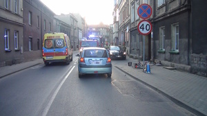 na zdjęciu droga, na której nieoznakowany radiowóz, karetka pogotowia oraz jasny pojazd biorący udział w zdarzeniu, obok policjanci i strażacy, a na chodniku przewrócony rower