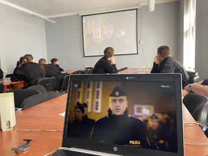 na zdjęciu na pierwszym planie laptop, na którego ekranie widoczny jest policjant, w drugim planie młodzież przy stole ogląda film wyświetlany na ekranie